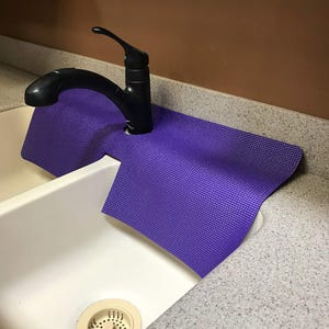 May include: A purple sink mat is placed under a black faucet in a white sink. The mat is designed to catch water and protect the countertop. The sink is made of white ceramic and has a drain with a beige strainer.
