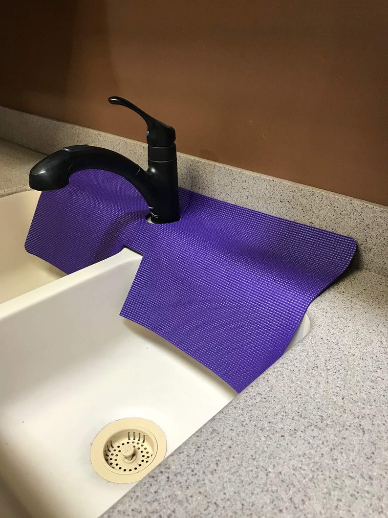 May include: A purple sink mat is placed under a black faucet in a white sink. The mat is designed to catch water and protect the countertop. The sink is made of white ceramic and has a drain with a beige strainer.