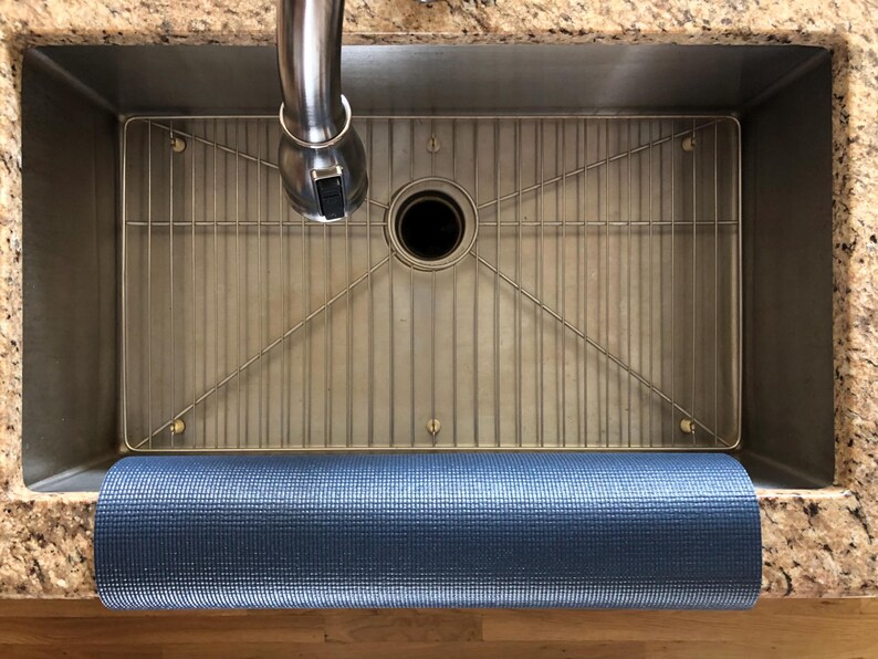 May include: A stainless steel kitchen sink with a metal grid and a rolled-up blue yoga mat. The sink is set into a granite countertop, with a chrome faucet above. The yoga mat is a textured, dark blue color.