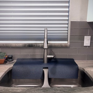 May include: A stainless steel kitchen sink with a modern faucet. A dark blue, textured mat sits over the sink's divider. The background includes a window with blinds and gray tile.