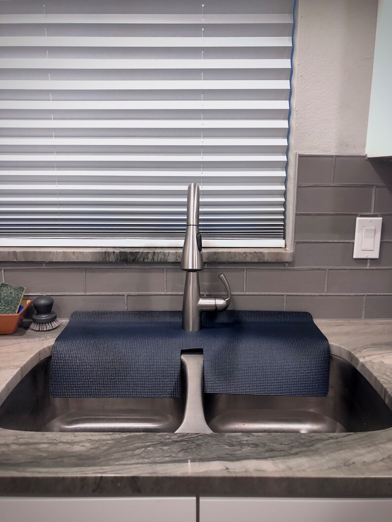 May include: A stainless steel kitchen sink with a modern faucet. A dark blue, textured mat sits over the sink's divider. The background includes a window with blinds and gray tile.