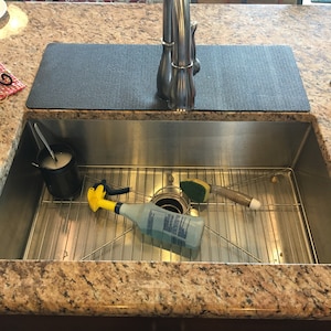 May include: A stainless steel kitchen sink with a chrome faucet. Inside the sink are cleaning supplies, including a yellow spray bottle, a sponge, and a container with soap. A black mat sits on the countertop behind the sink.