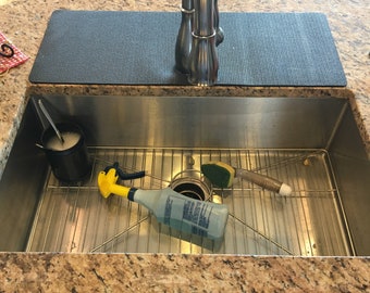 May include: A stainless steel kitchen sink with a chrome faucet. Inside the sink are cleaning supplies, including a yellow spray bottle, a sponge, and a container with soap. A black mat sits on the countertop behind the sink.