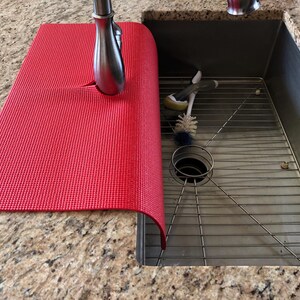 May include: A red textured sink mat partially covering a stainless steel sink. The mat is positioned under a silver faucet. A metal sink rack and cleaning brushes are visible inside the sink.