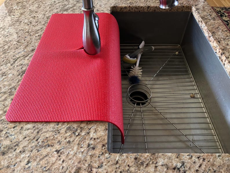 May include: A red textured sink mat partially covering a stainless steel sink. The mat is positioned under a silver faucet. A metal sink rack and cleaning brushes are visible inside the sink.