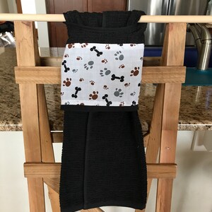 May include: A black kitchen towel with a white band featuring paw prints and bones in brown, grey, and black. The towel is hanging on a wooden rack, showcasing a pet-themed design.