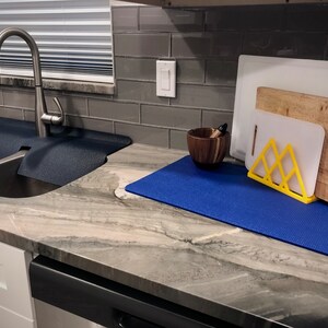 May include: A kitchen scene featuring a stainless steel sink with a dark blue mat, a faucet, and a countertop with cutting boards, a wooden mortar and pestle, and a yellow stand. The countertop is a light gray with darker veining.