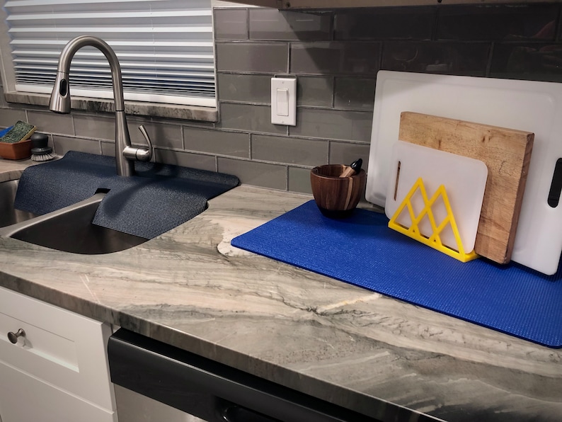 May include: A kitchen scene featuring a stainless steel sink with a dark blue mat, a faucet, and a countertop with cutting boards, a wooden mortar and pestle, and a yellow stand. The countertop is a light gray with darker veining.
