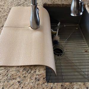 May include: A beige sink mat with a textured surface, partially covering a stainless steel sink. The mat is positioned near a chrome faucet. A metal sink rack and cleaning brushes are visible inside the sink.
