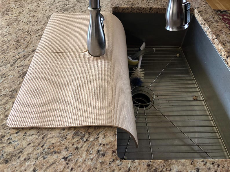 May include: A beige sink mat with a textured surface, partially covering a stainless steel sink. The mat is positioned near a chrome faucet. A metal sink rack and cleaning brushes are visible inside the sink.
