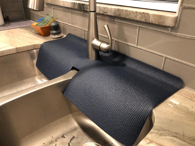 May include: A navy blue sink mat with a textured surface, designed to fit around a faucet. The mat is placed on a granite countertop next to a stainless steel sink. A dish brush and sponge are visible in the background.