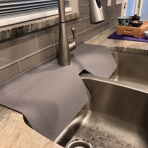 May include: A gray sink mat with a textured surface, designed to fit around a kitchen faucet and protect the countertop. The mat is placed over a stainless steel sink. A tea set is visible in the background.