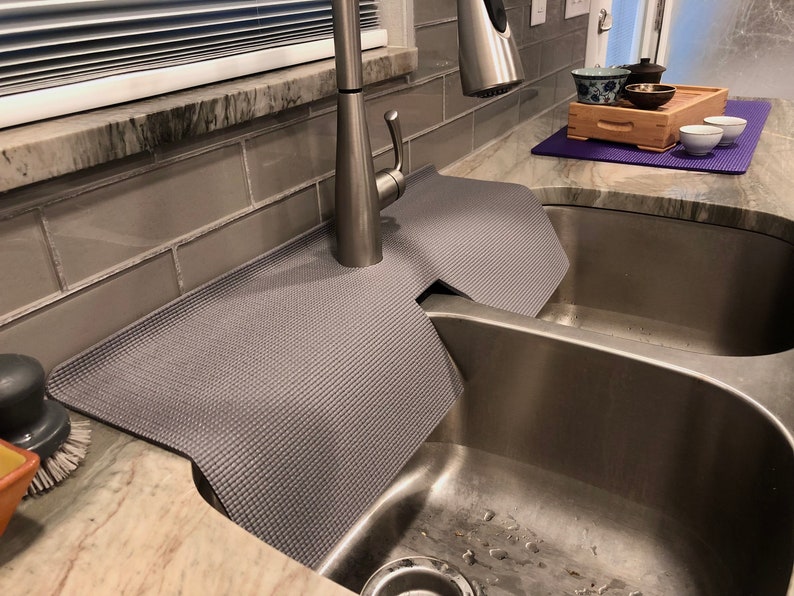 May include: A gray sink mat with a textured surface, designed to fit around a kitchen faucet and protect the countertop. The mat is placed over a stainless steel sink. A tea set is visible in the background.