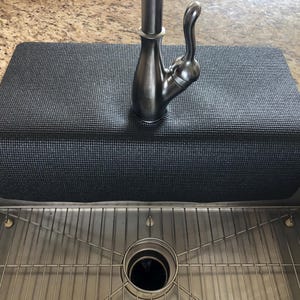 May include: A black sink mat with a textured surface, designed to fit over the edge of a kitchen sink. The mat is positioned under a silver faucet and above a stainless steel sink with a metal grid.