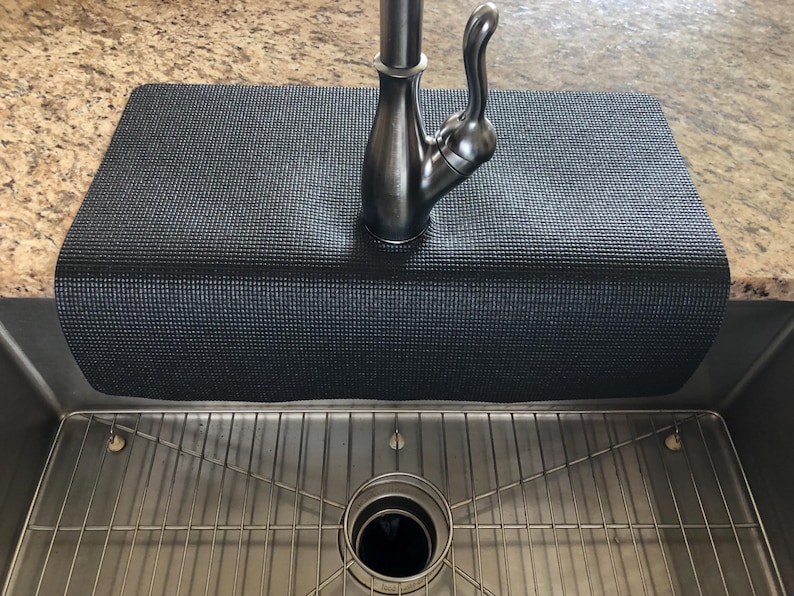 May include: A black sink mat with a textured surface, designed to fit over the edge of a kitchen sink. The mat is positioned under a silver faucet and above a stainless steel sink with a metal grid.