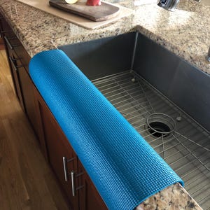 May include: A rolled-up, textured, bright blue kitchen sink mat rests on a granite countertop. The mat is positioned next to a stainless steel sink. A wooden cutting board with sliced apples and a knife is visible.
