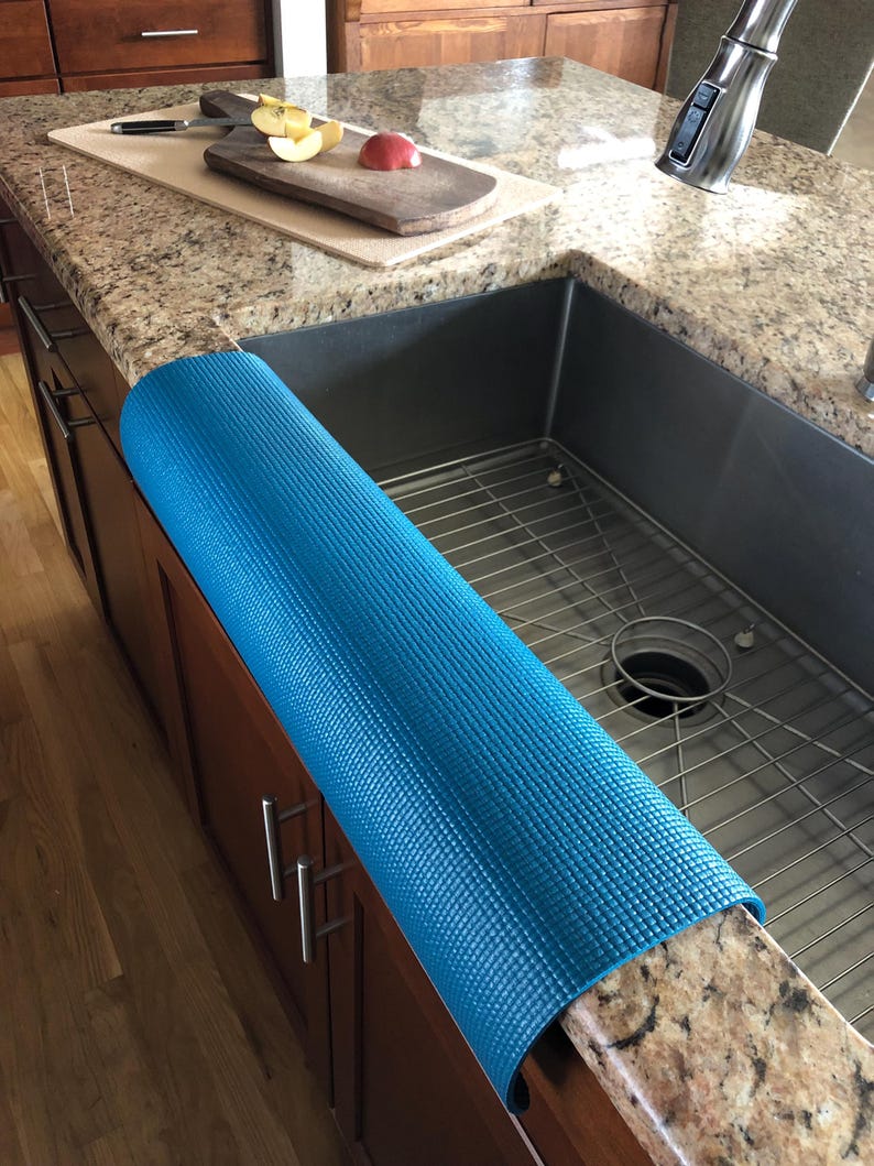 May include: A rolled-up, textured, bright blue kitchen sink mat rests on a granite countertop. The mat is positioned next to a stainless steel sink. A wooden cutting board with sliced apples and a knife is visible.