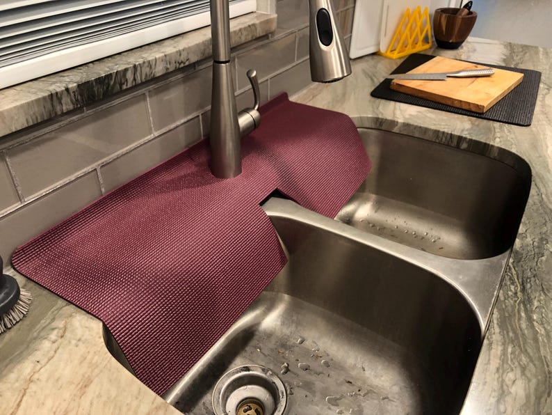 May include: A burgundy sink mat is draped over a stainless steel double sink. The mat is made of a textured material and is designed to protect the countertop from water damage. A stainless steel tap is visible in the background.