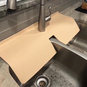 May include: A beige sink mat with a textured surface, designed to fit around a faucet. The mat is placed over a stainless steel sink, providing a protective barrier against water and potential scratches. The mat's neutral color complements the sink's metallic finish.