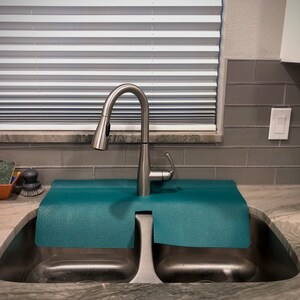 May include: A modern stainless steel kitchen tap with a pull-down spray head, positioned above a double sink. A teal mat sits between the tap and the sink basins. The background features blinds and grey tiles.