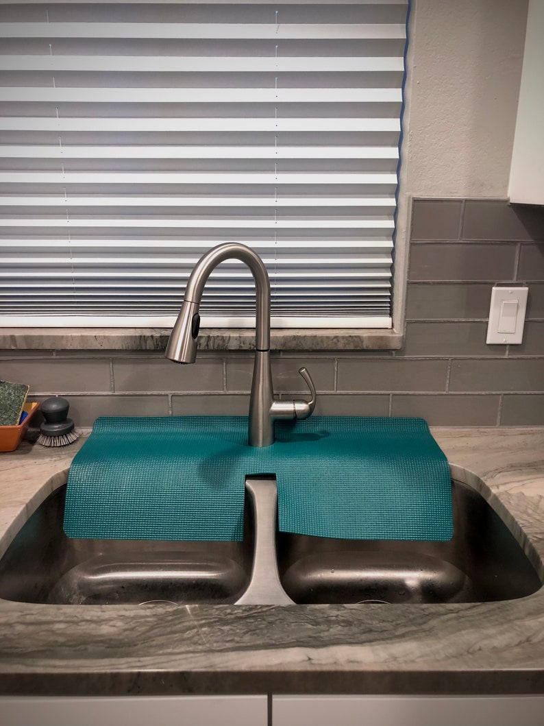 May include: A modern stainless steel kitchen tap with a pull-down spray head, positioned above a double sink. A teal mat sits between the tap and the sink basins. The background features blinds and grey tiles.