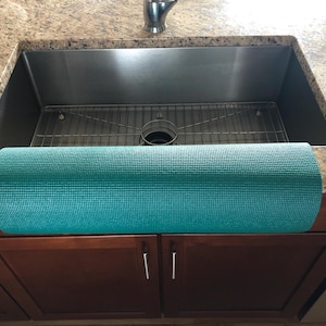 May include: A rolled-up teal yoga mat rests on a granite countertop in front of a stainless steel sink. The mat is textured and appears to be made of a durable material. The kitchen cabinets below are a dark brown color.