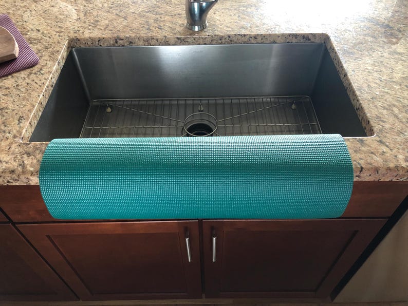 May include: A rolled-up teal yoga mat rests on a granite countertop in front of a stainless steel sink. The mat is textured and appears to be made of a durable material. The kitchen cabinets below are a dark brown color.