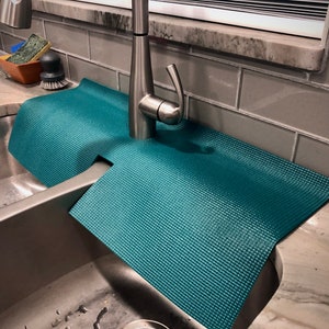 May include: Teal sink mat with a textured surface, designed to fit around a tap. The mat is placed in a stainless steel sink, with a tap and cleaning supplies visible in the background. The mat is made of a flexible material.