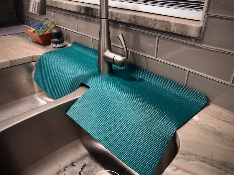 May include: Teal sink mat with a textured surface, designed to fit around a tap. The mat is placed in a stainless steel sink, with a tap and cleaning supplies visible in the background. The mat is made of a flexible material.