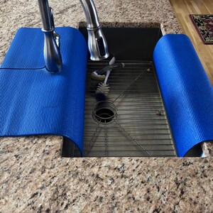 May include: A blue sink mat is draped over the edge of a stainless steel sink. The mat is made of a textured material and is designed to protect the countertop from water damage. A chrome faucet is visible in the background.