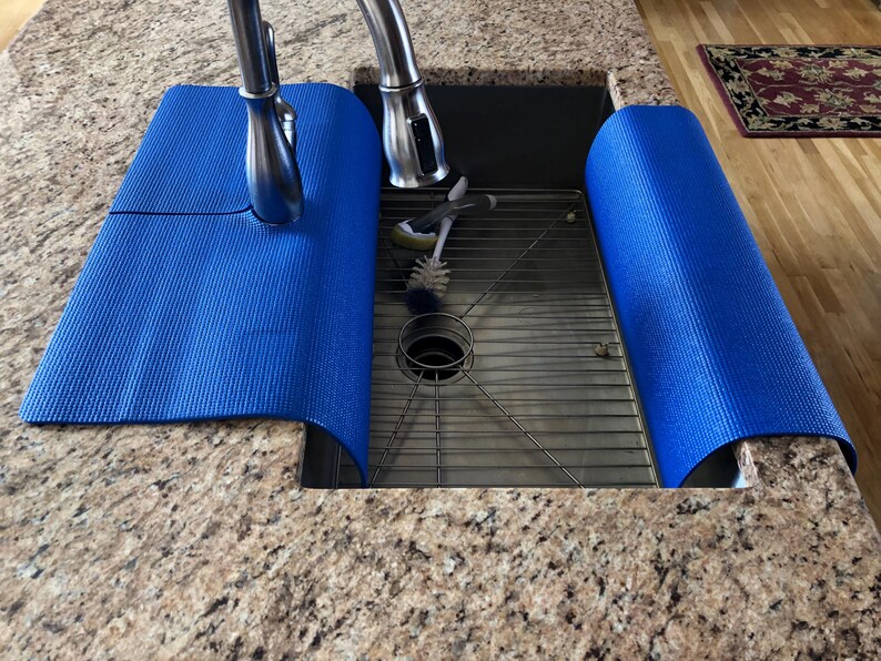 May include: A blue sink mat is draped over the edge of a stainless steel sink. The mat is made of a textured material and is designed to protect the countertop from water damage. A chrome faucet is visible in the background.