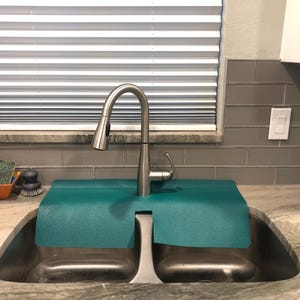 May include: A stainless steel kitchen faucet with a pull-down sprayer is installed over a double sink. A teal-colored mat sits between the faucet and the sink basins. The sink is set into a granite countertop.
