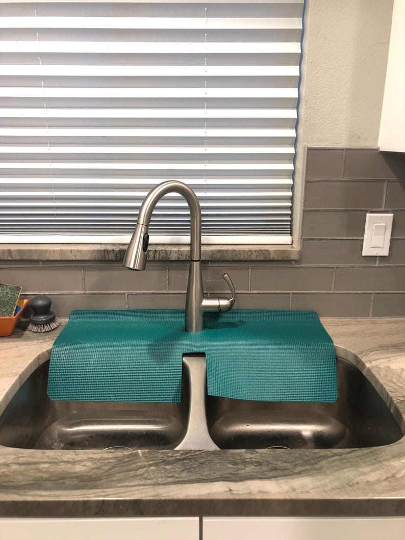 May include: A stainless steel kitchen faucet with a pull-down sprayer is installed over a double sink. A teal-colored mat sits between the faucet and the sink basins. The sink is set into a granite countertop.