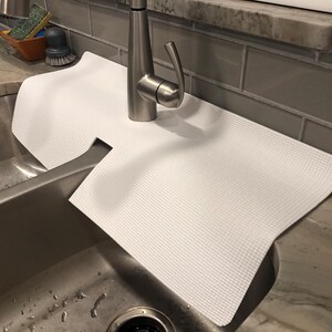 May include: A white, textured sink mat is placed over a stainless steel sink. The mat has a cutout for the faucet and is designed to protect the sink's surface. The faucet is made of brushed nickel.