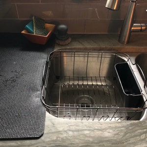 May include: A stainless steel kitchen sink with a wire dish rack and a black cutting board. A small orange bowl holds sponges and a scrub brush. The faucet is chrome.
