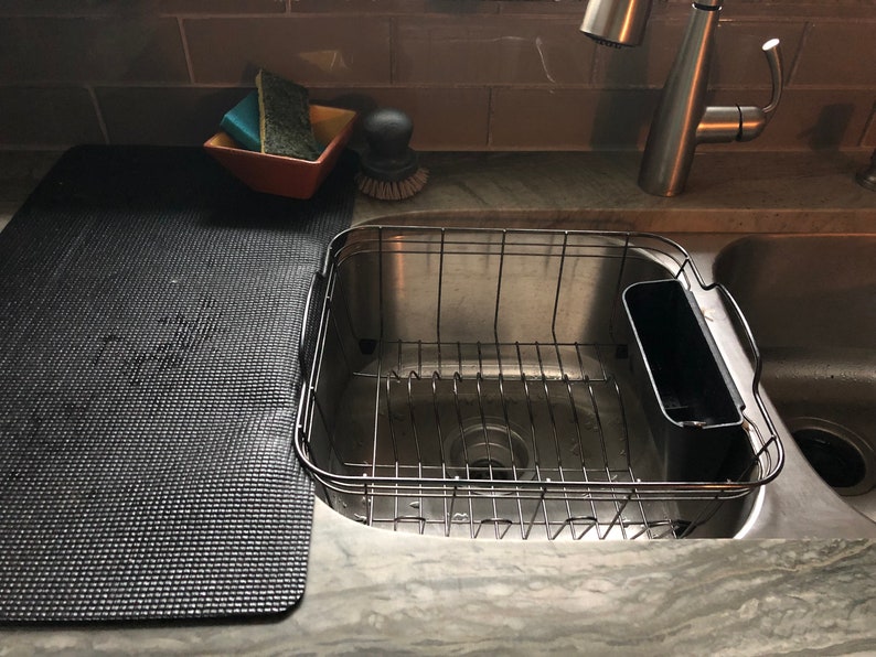 May include: A stainless steel kitchen sink with a wire dish rack and a black cutting board. A small orange bowl holds sponges and a scrub brush. The faucet is chrome.