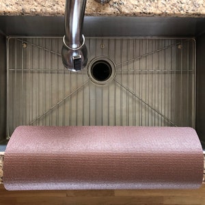 May include: A rolled-up, textured, pink yoga mat rests in a stainless steel kitchen sink. The sink has a metal grid and a chrome faucet. The countertop is granite.