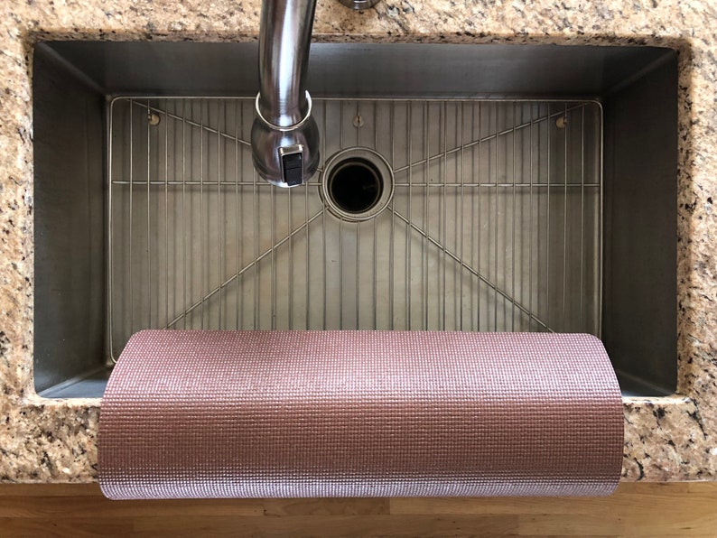 May include: A rolled-up, textured, pink yoga mat rests in a stainless steel kitchen sink. The sink has a metal grid and a chrome faucet. The countertop is granite.
