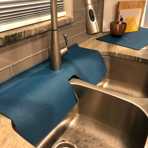 May include: A blue sink mat is placed around a stainless steel faucet and sink. The mat is made of a textured material and is designed to protect the countertop from water damage. Additional kitchen accessories are visible in the background.