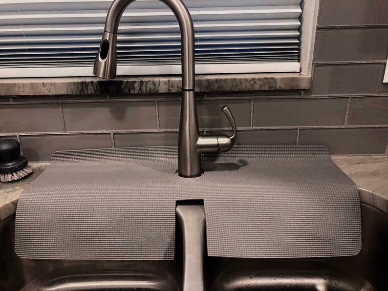 May include: A gray sink mat with a textured surface is placed under a stainless steel kitchen faucet. The faucet has a pull-down sprayer and a curved spout. The sink is stainless steel, and the background includes gray tile and a window.