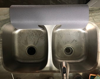 Könnte beinhalten: Eine aufgerollte, hellgraue, strukturierte Spülbeckenmatte liegt über einem Doppelwaschbecken aus Edelstahl. Die Matte besteht aus einem flexiblen Material und soll die Oberfläche des Waschbeckens schützen. Das Waschbecken hat zwei Becken und eine Chromarmatur.