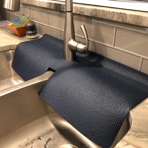May include: A navy blue sink mat with a textured surface, designed to fit around a tap. The mat is placed on a granite countertop next to a stainless steel sink. The mat is designed to catch water and protect the countertop.