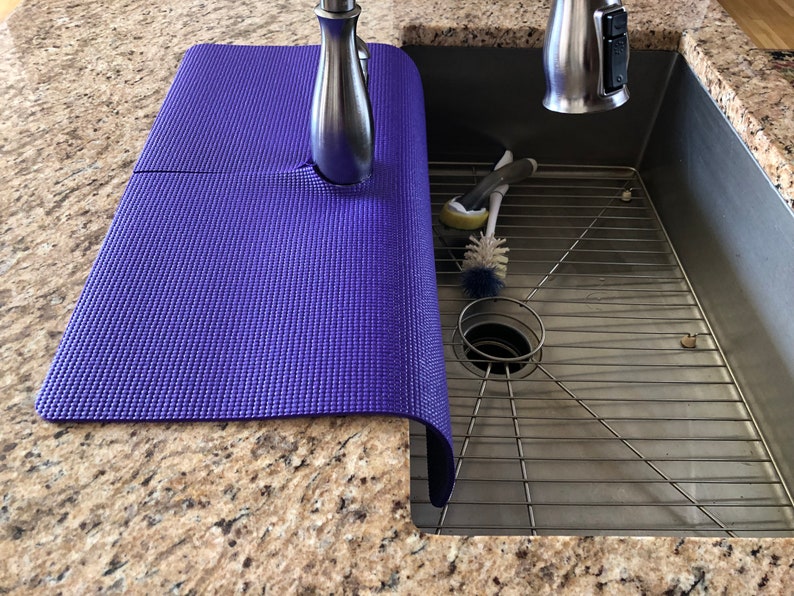 May include: A purple sink mat is draped over the edge of a stainless steel sink. The mat is made of a textured material and is positioned under a chrome faucet. Cleaning brushes are visible inside the sink.