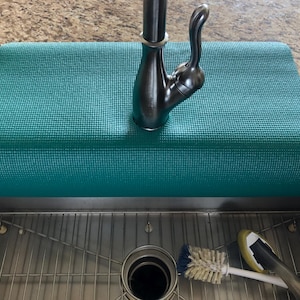 May include: A teal-colored, rectangular sink mat rests beneath a dark metal faucet. The mat is textured and sits above a stainless steel sink with a metal grate. A scrub brush and a sponge are visible.