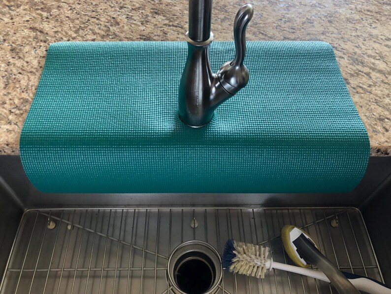May include: A teal-colored, rectangular sink mat rests beneath a dark metal faucet. The mat is textured and sits above a stainless steel sink with a metal grate. A scrub brush and a sponge are visible.