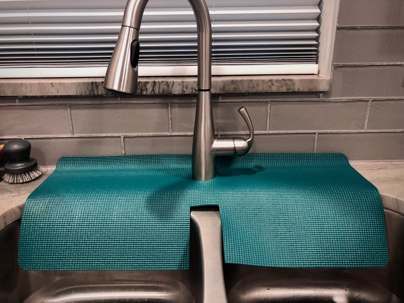 May include: A teal sink mat is positioned under a stainless steel kitchen tap. The mat, with a textured surface, is designed to protect the worktop from water. The tap features a pull-down spray function.