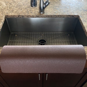 May include: A stainless steel kitchen sink with a metal grid and a rolled-up, textured, brown mat. The sink is set into a granite countertop, with a faucet and cabinet visible.