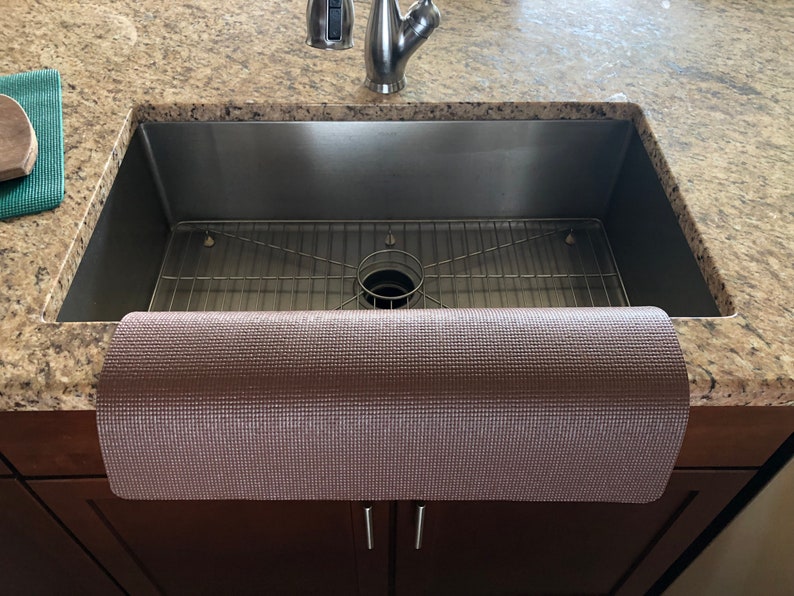 May include: A stainless steel kitchen sink with a metal grid and a rolled-up, textured, brown mat. The sink is set into a granite countertop, with a faucet and cabinet visible.