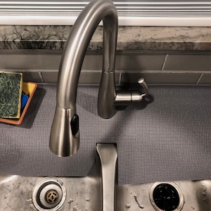 May include: A stainless steel kitchen faucet with a pull-down sprayer is positioned over a stainless steel sink. A gray textured mat sits beneath the faucet. A sponge and a blue cloth rest on a yellow and brown dish. The background includes a window and gray tile.