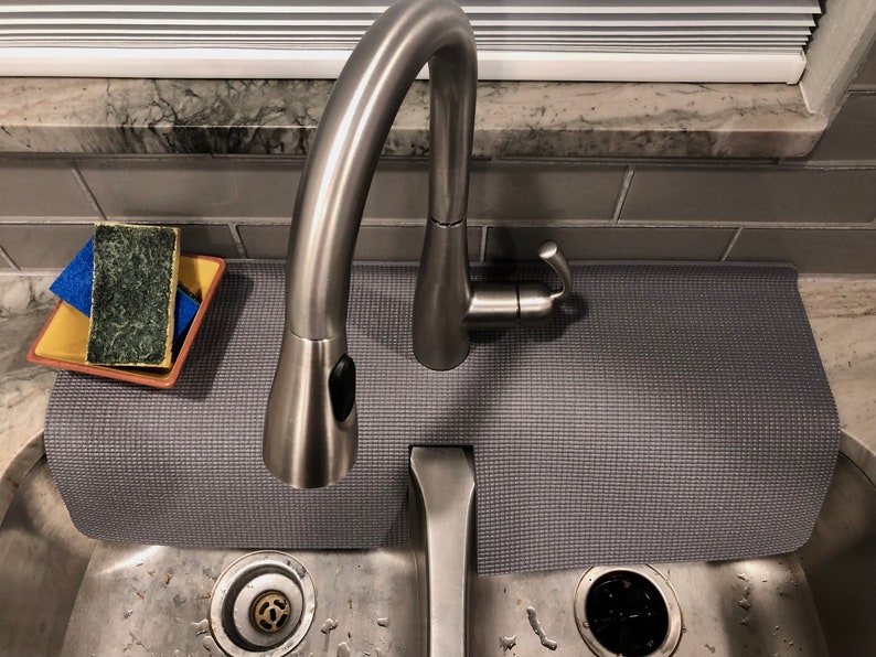 May include: A stainless steel kitchen faucet with a pull-down sprayer is positioned over a stainless steel sink. A gray textured mat sits beneath the faucet. A sponge and a blue cloth rest on a yellow and brown dish. The background includes a window and gray tile.